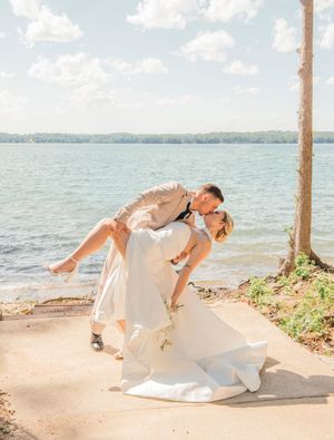 A romantic wedding couple shares a passionate dip kiss on a lakeside path with sunlit water and trees in the background.