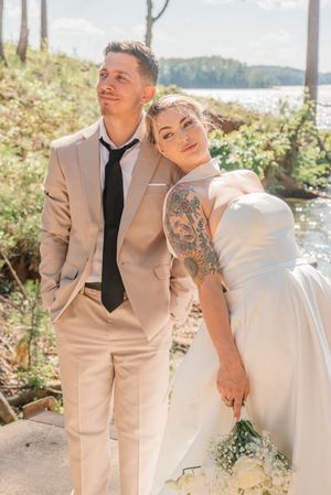 A couple in wedding attire poses outdoors with scenic water and forest views in the background on a sunny day.