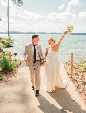 A newlywed couple celebrates their wedding day walking along a sunny lakeside path in summer attire.