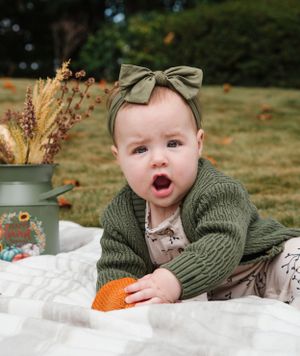 A baby in an olive green sweater and matching bow headband lies on a white blanket with an orange toy.
