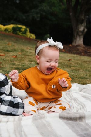 Joyful baby in orange sweater laughs while sitting on white blanket outdoors.