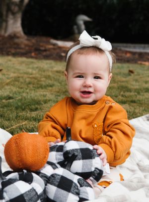 Baby in orange sweater sits on blanket with pumpkin and plaid decorations.