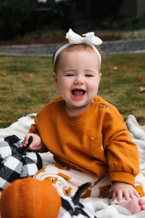 Laughing baby wearing orange sweater and white bow sits with fall decorations.