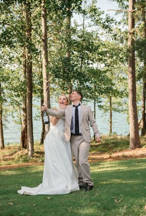 A joyful couple walks together among tall trees by a lake on their wedding day in natural outdoor setting.