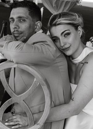 Black and white portrait of a couple in formal attire hugging and smiling while sitting in an ornate chair.