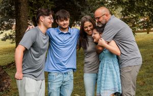 A family of five wearing casual clothing embraces while standing together outdoors in a park setting.