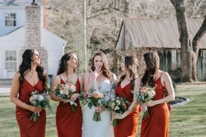 A bridal party in red dresses holding bouquets poses with the bride in white on a sunny day.