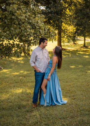 A couple in a grassy park setting during golden hour, with the person in a flowing blue dress and white shirt.
