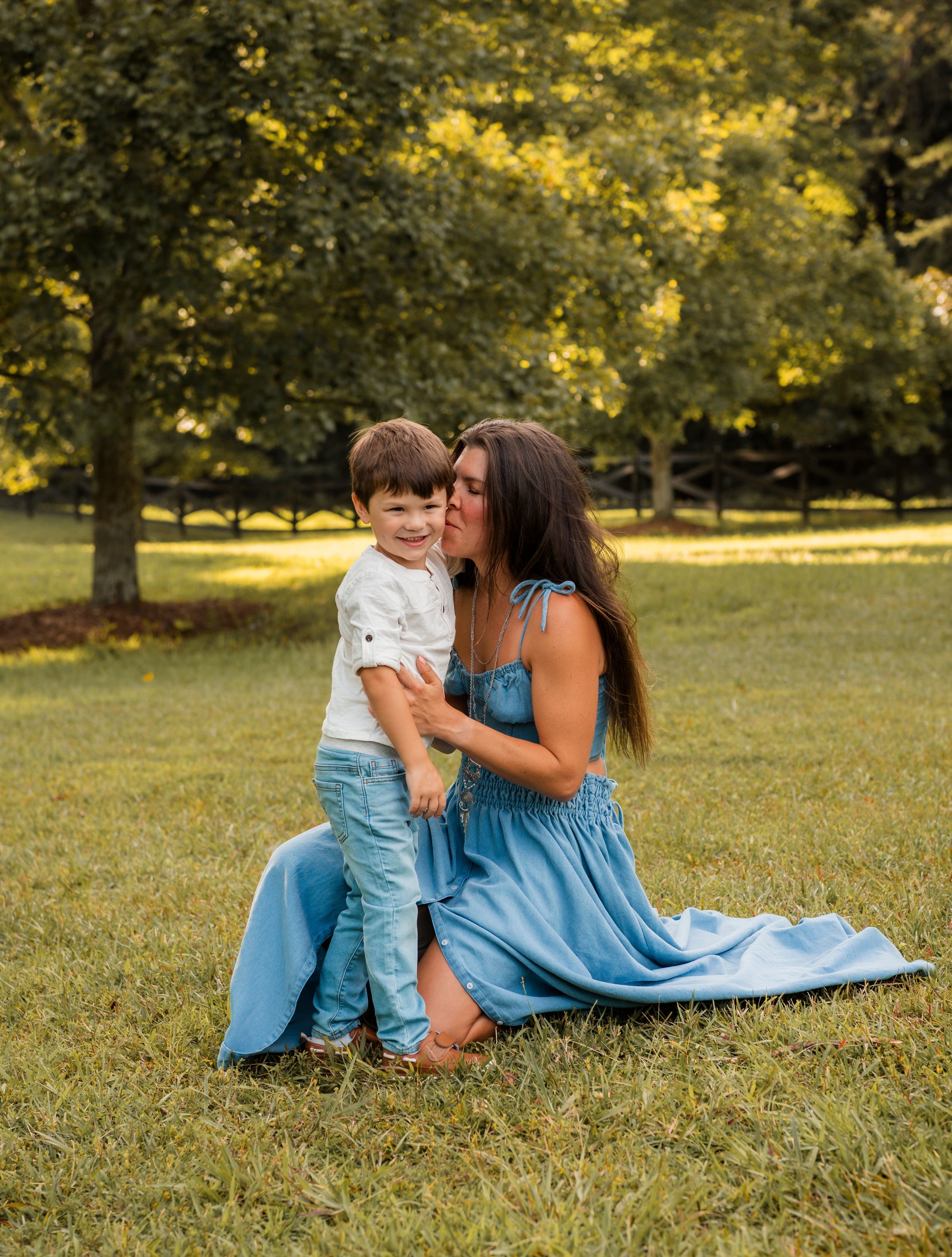 A mother and young child in matching blue outfits share tender moments while sitting on grass in a sunlit park.
