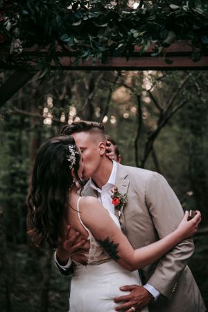 A romantic wedding couple shares a kiss under trees with dreamy bokeh lighting in the background.