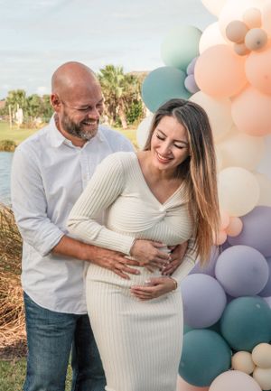 Expecting couple sharing tender moment against pastel balloon backdrop outdoors.