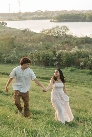Couple holds hands while walking through a grassy hillside overlooking a serene lake at sunset.