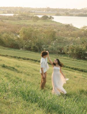 Romantic couple walks hand in hand on a green hillside with water views in golden light.