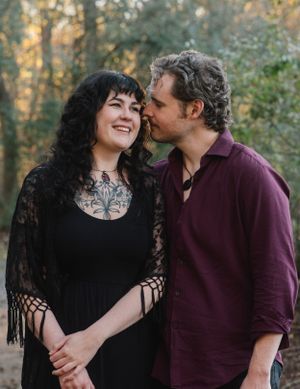 Couple shares intimate moment outdoors wearing black and burgundy against forest background.