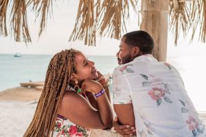 A couple shares a playful moment on a beach under a thatched umbrella.