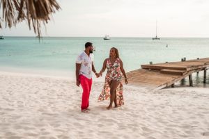 A couple walks hand in hand on a white sandy beach with a wooden pier in the background.