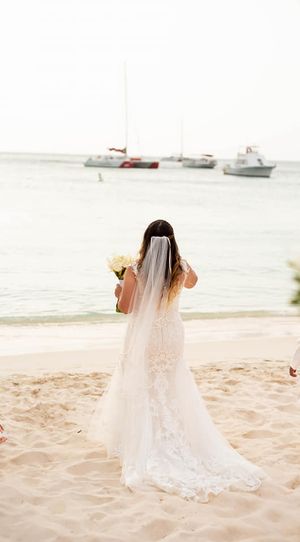 A bride in a flowing lace wedding dress stands on a beach looking at sailboats on the horizon.