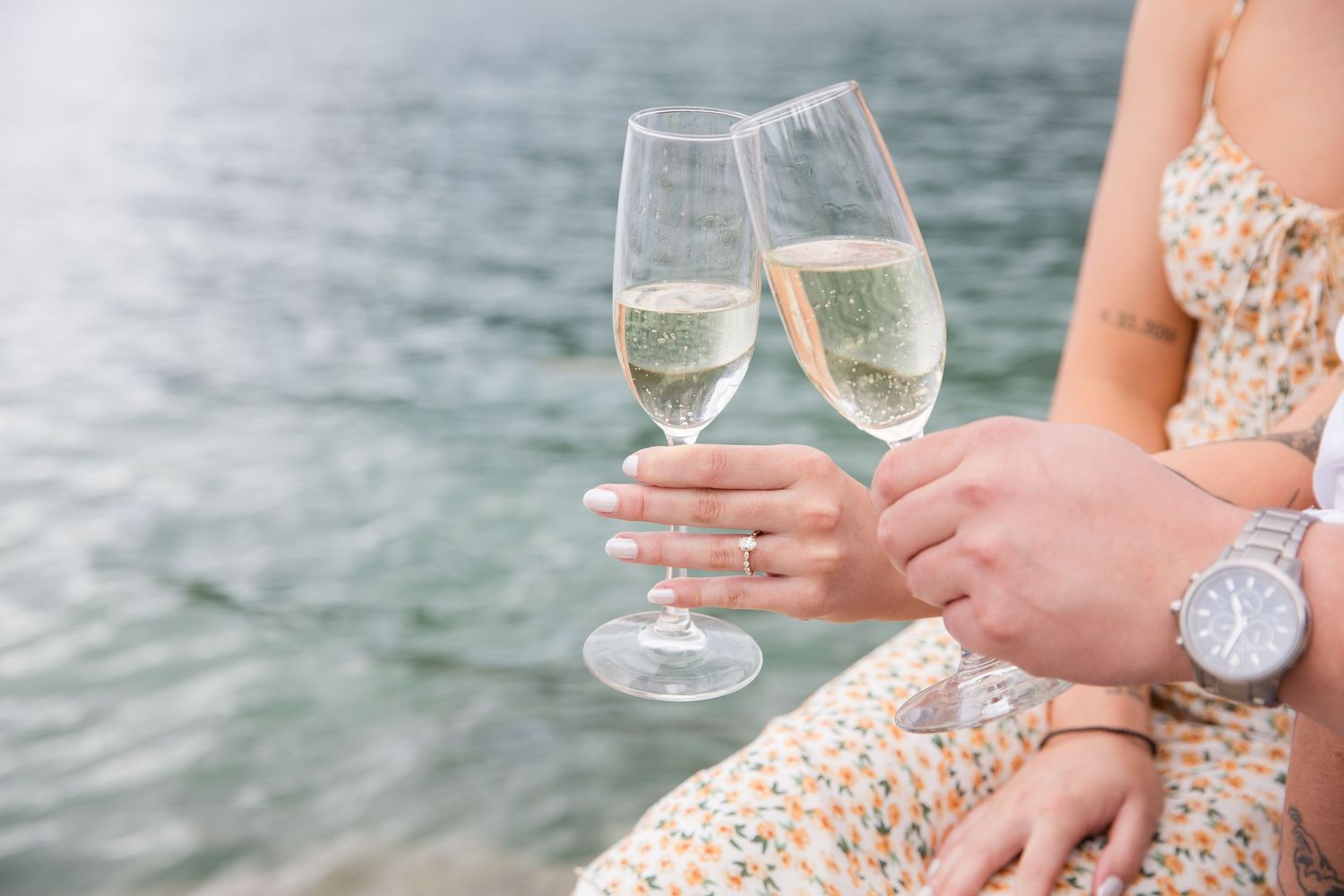 Two champagne glasses clink together against water backdrop with floral dress and jewelry visible.