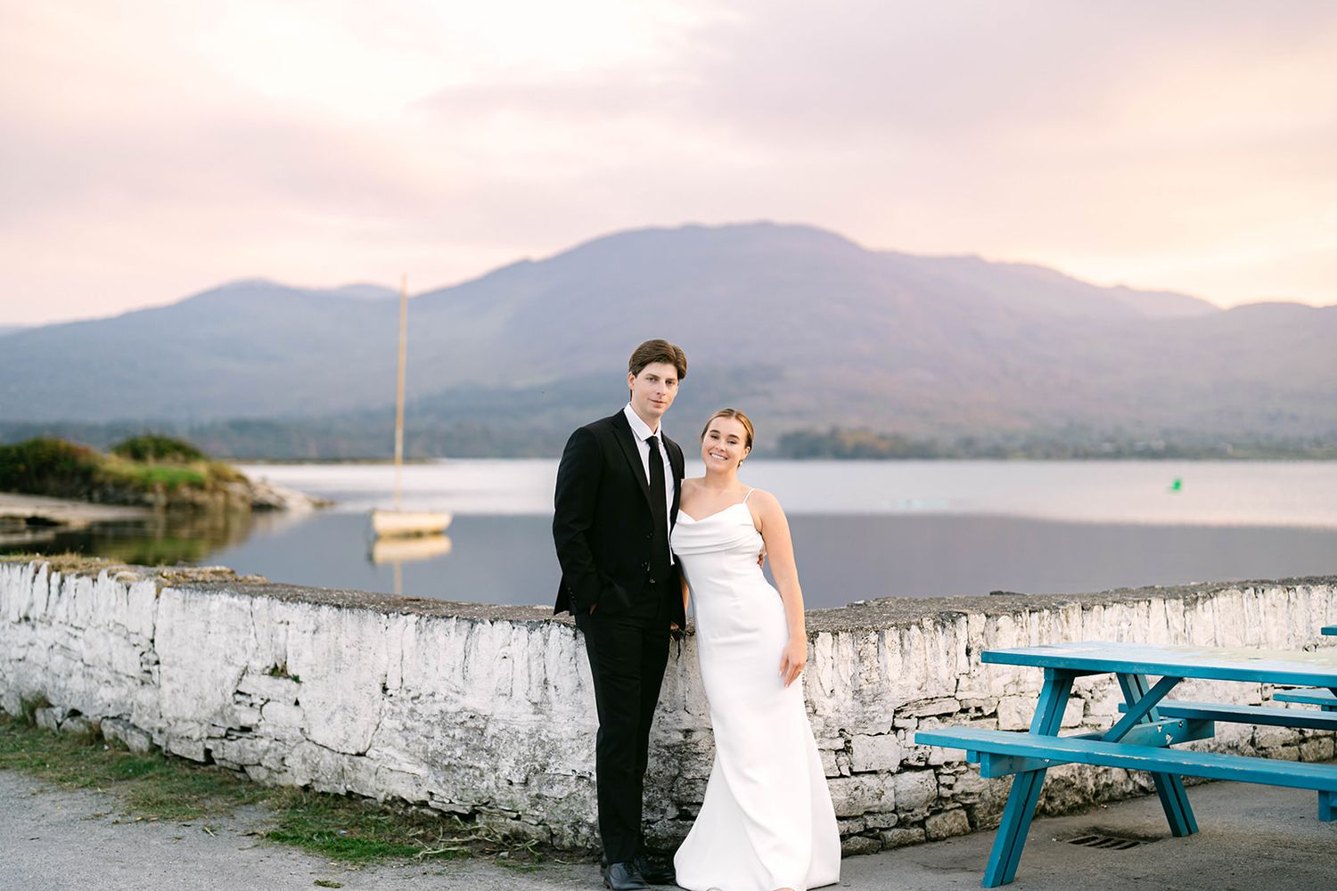 Newlyweds pose by a serene lake with mountains and a blue picnic table in the scene.