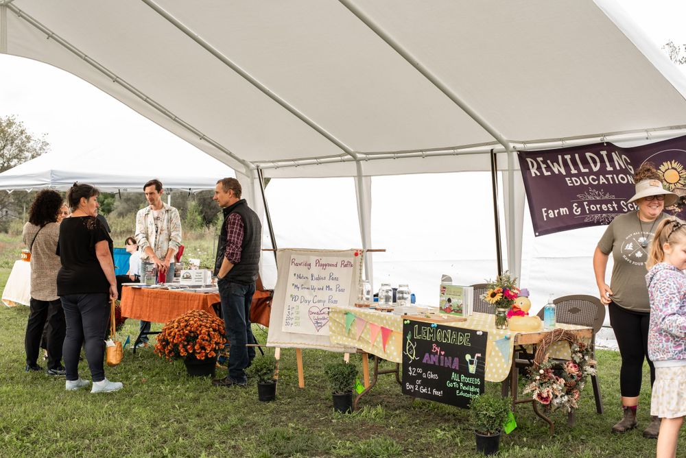 Outdoor market vendors and customers gather under white tent with fall decorations.