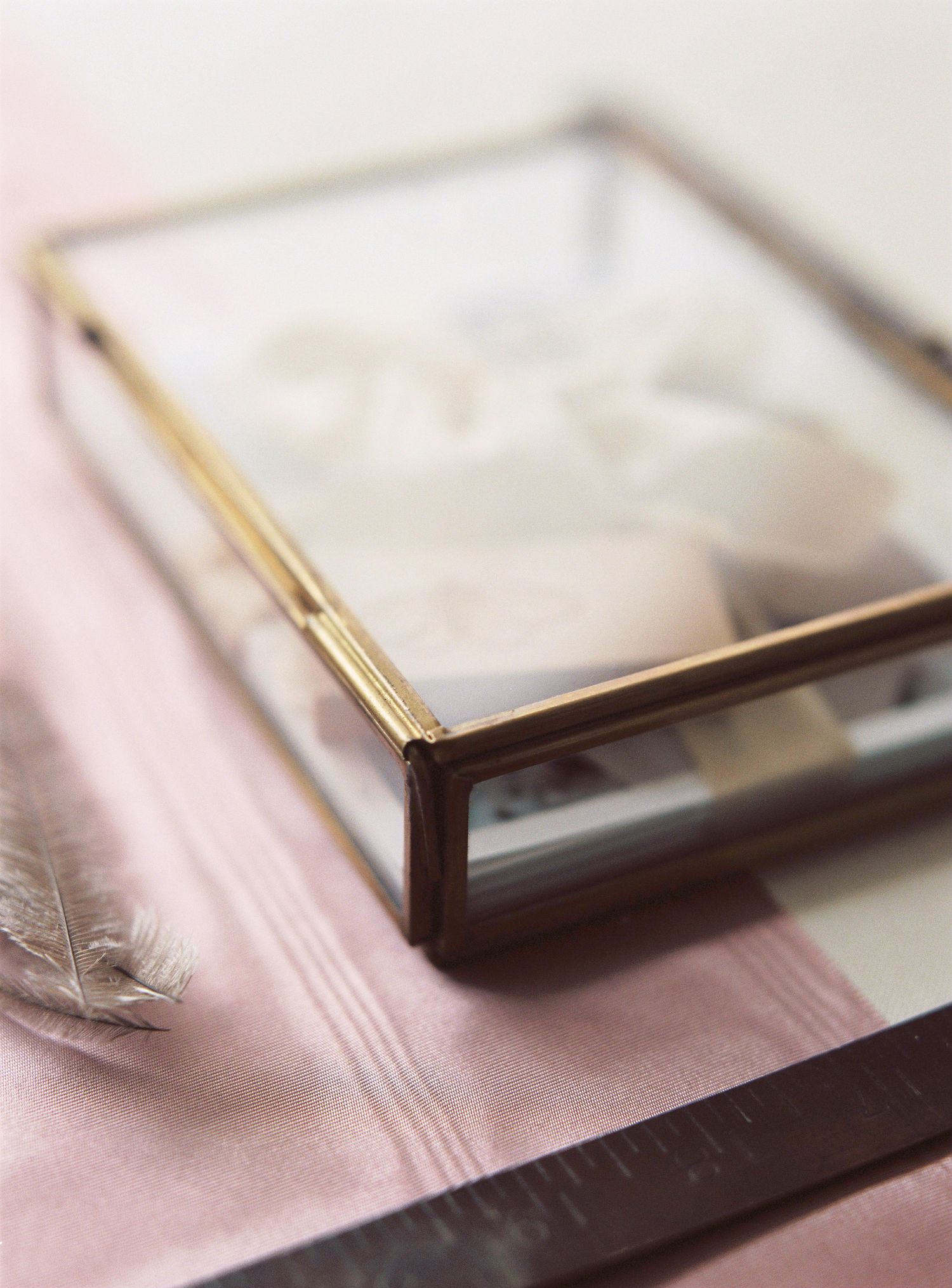 A delicate glass and brass display box on pink fabric with a white feather beside it.