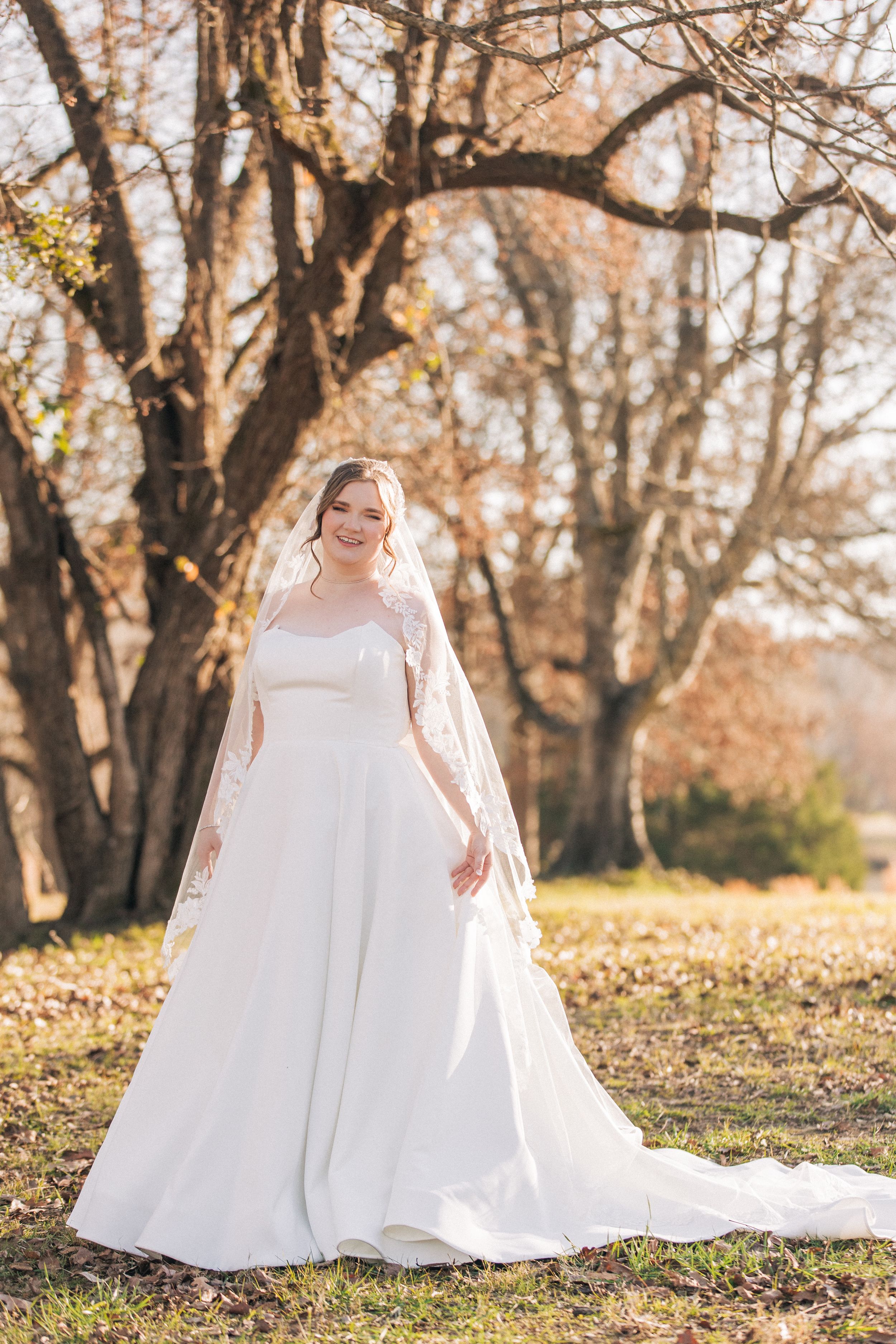 A bride in a flowing white wedding dress sits gracefully among autumn trees during an outdoor photo session.