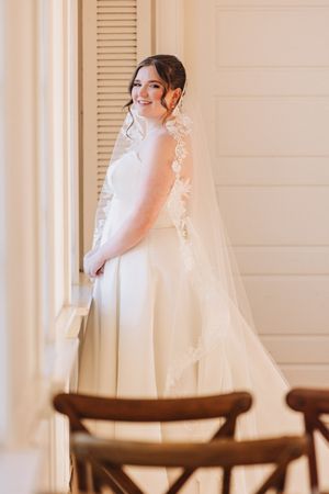 A bride in a white wedding gown with long veil poses delicately in a room with wooden chairs.