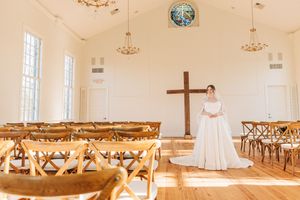 Panoramic view of a bright white church interior with wooden pews, chandeliers, and a cross mounted on the wall.