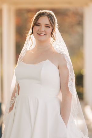 Elegant bride in simple white sleeveless wedding dress and delicate veil standing in soft natural light.