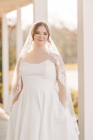 Elegant bride in simple white wedding dress and delicate lace veil poses with soft natural lighting in outdoor setting.