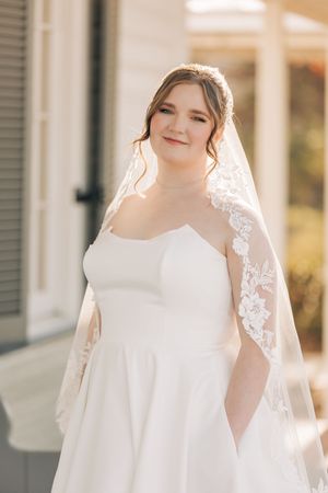 A bride in a classic white wedding gown and delicate lace veil stands in soft sunlight against white shutters.