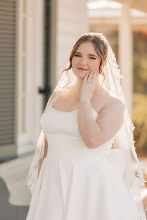 A bride in a flowing white dress and veil stands on a sunlit porch, touching her face gently with a joyful expression.