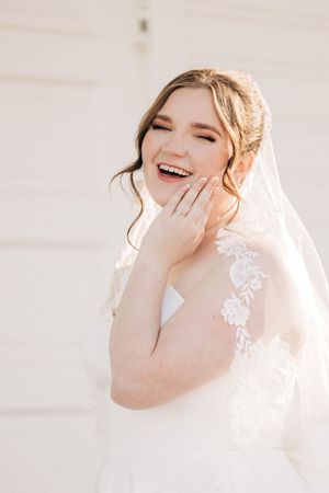 Glowing bride in white lace dress and veil laughing joyfully against light background.