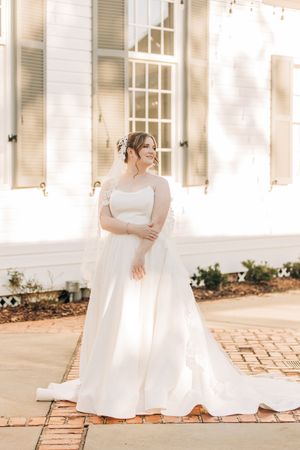 Bride in flowing off-shoulder wedding gown poses elegantly on brick pathway beside white building with classic shutters.