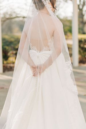 Back view of a bride in an elegant white wedding gown with sheer veil showing delicate lace details in an outdoor setting.
