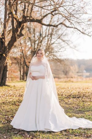 A bride in a stunning white ballgown wedding dress and cathedral veil stands gracefully among autumn trees.