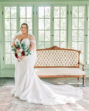 A bride wearing a fitted lace gown holds colorful bouquet while standing in front of vintage settee in green room.