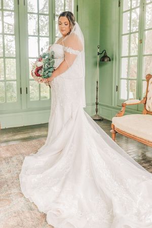 A bride in an off-shoulder lace wedding gown with long train poses in a green sunlit room with French doors.
