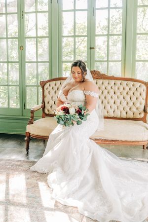 Elegant bride in off-shoulder white gown sits gracefully on vintage tufted sofa near sunlit windows holding floral bouquet.