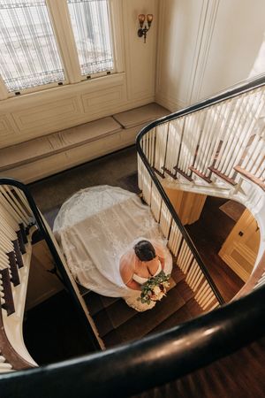 Aerial view of a curved staircase with elegant black railing and a flowing white wedding dress from above.