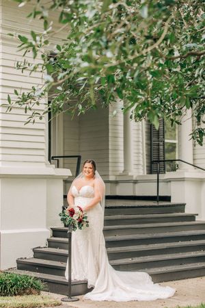 A bride in a white wedding dress poses on stone steps outside a classic white house with black shutters and overhanging trees.