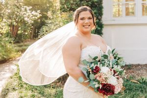 Person in white lace wedding dress holding beautiful bouquet with burgundy and blush flowers and flowing veil outdoors.