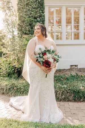 Beautiful bride wearing lace wedding dress and cape stands outdoors holding burgundy and blush floral bouquet.