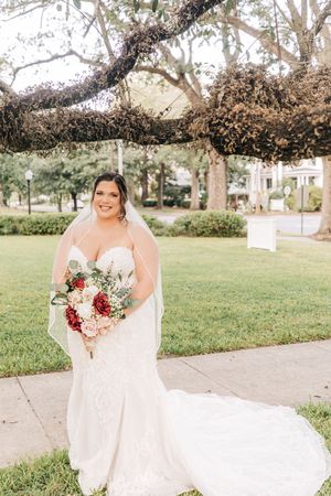 A bride in a white lace mermaid wedding gown holds a vibrant red and pink bouquet while standing in a garden setting.