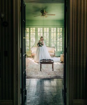 Wedding couple stands in sunlit interior of rustic building with window-lit reflections on wooden floor.