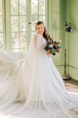 A person in a romantic white cape gown holds a colorful wedding bouquet near a bright window with sage green walls.