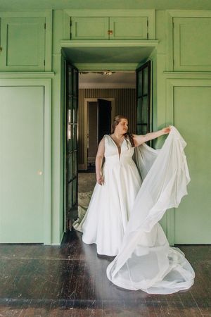 A person in a flowing white wedding dress stands in front of mint green cabinets while holding their veil.