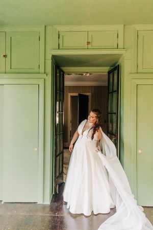 A person in an elegant white wedding gown poses in a doorway with sage green walls and built-in cabinets.
