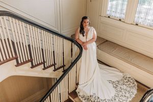 A bride in a white wedding dress descends a curved staircase in an elegant light-colored interior setting.
