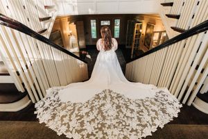 Elegant bride in white wedding gown with ornate lace train stands atop grand staircase viewed from behind.